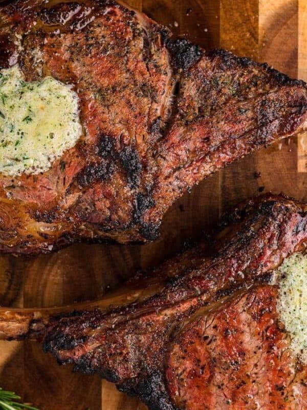 Two grilled tomahawk steaks topped with herb butter are on a wooden cutting board. Fresh rosemary sprigs are placed nearby for garnish. The steaks have a charred, juicy appearance.