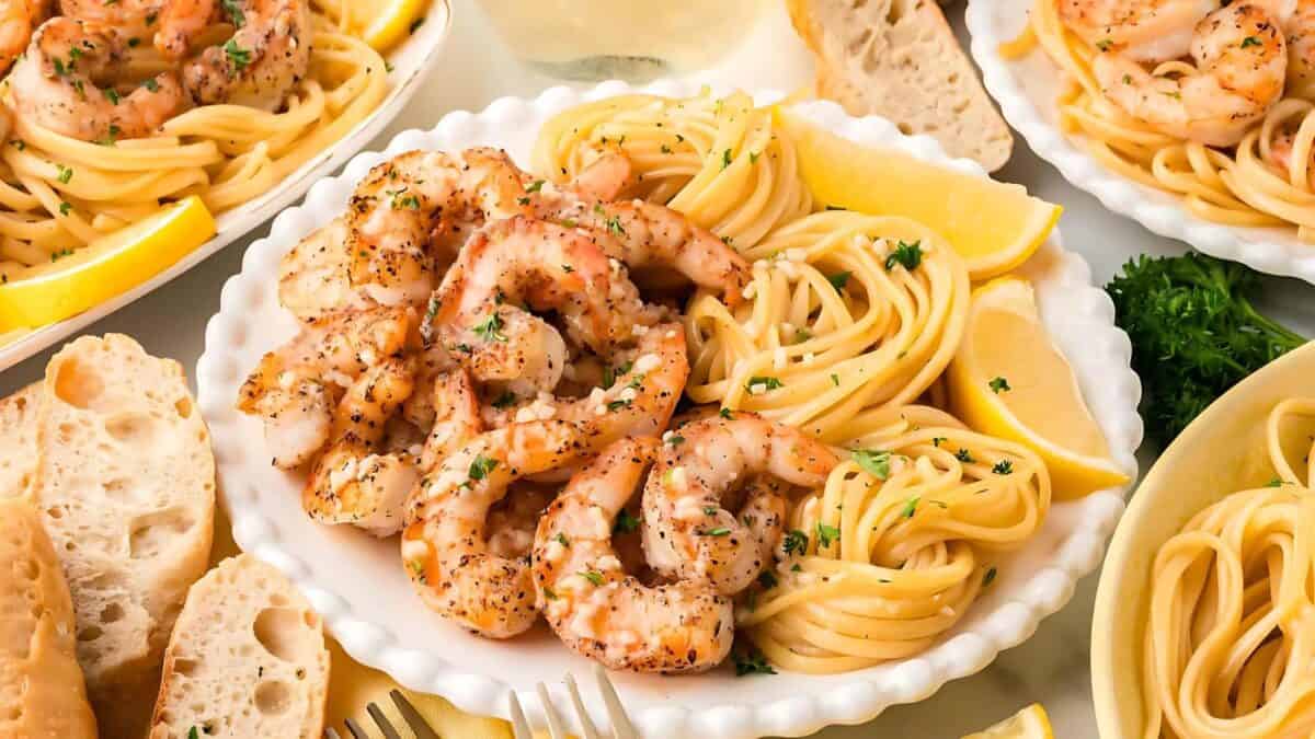 The image shows several plates of spaghetti with garlic shrimp. The shrimp are cooked and seasoned, and the spaghetti is tossed in a light sauce. Lemon wedges and parsley are used as garnishes. Slices of bread are also visible. The plates are white with scalloped edges. The background is a marble surface.