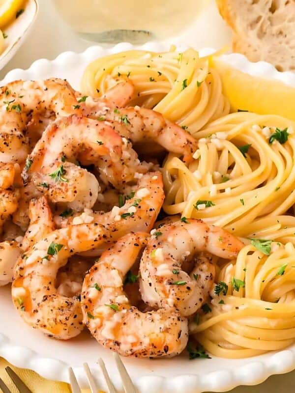 The image shows several plates of spaghetti with garlic shrimp. The shrimp are cooked and seasoned, and the spaghetti is tossed in a light sauce. Lemon wedges and parsley are used as garnishes. Slices of bread are also visible. The plates are white with scalloped edges. The background is a marble surface.