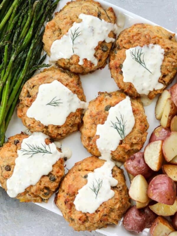 A platter with grilled asparagus, five salmon patties topped with white sauce and dill, and roasted red potatoes. A lemon wedge and a small bowl of sauce with a spoon are visible on the side.