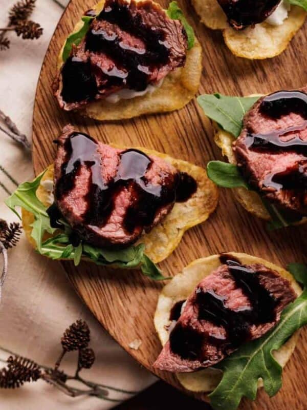 The image shows a wooden board of appetizers. Each appetizer consists of a potato chip topped with a piece of seared steak, a dollop of what appears to be goat cheese, arugula, and a drizzle of balsamic glaze. The board is rustic and the overall presentation is elegant and suggests a sophisticated hors d'oeuvre. A section of antler is visible in the background.