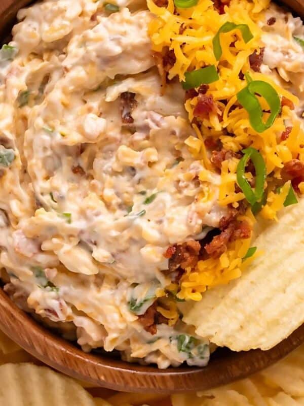 The image shows a wooden bowl of creamy bacon and cheese dip. The dip is topped with shredded cheddar cheese, crumbled bacon, and chopped green onions. Potato chips surround the bowl.