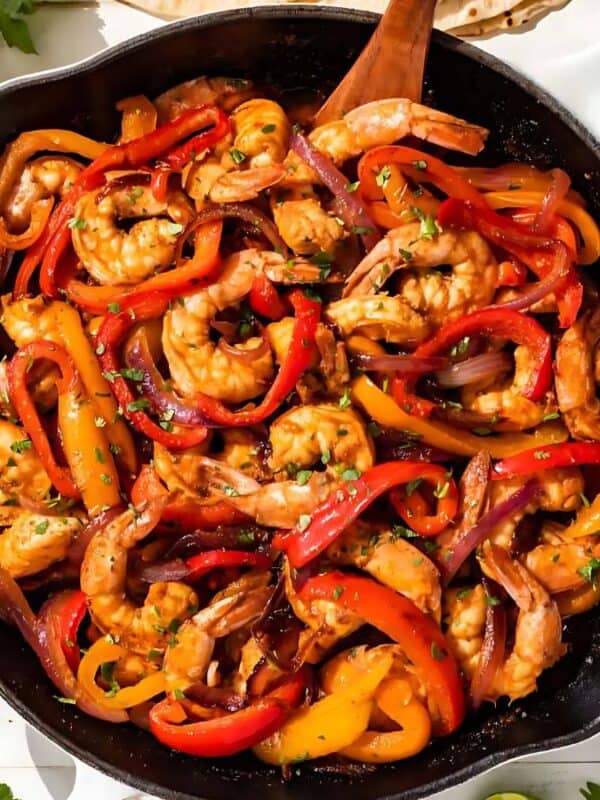 A skillet filled with cooked shrimp, red and orange bell peppers, onions, and seasoning. The dish is surrounded by a striped cloth, sliced avocado, fresh cilantro, lime wedges, sour cream, and a salsa bowl.