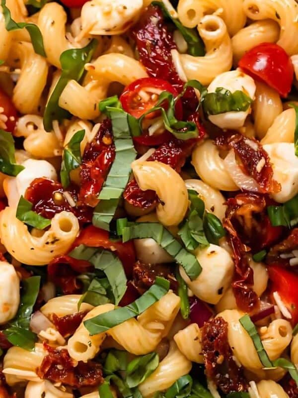 A vibrant pasta salad features spiral pasta, cherry tomatoes, sun-dried tomatoes, fresh basil, and mozzarella balls. The dish is garnished with sliced basil leaves, creating a colorful and appetizing presentation.