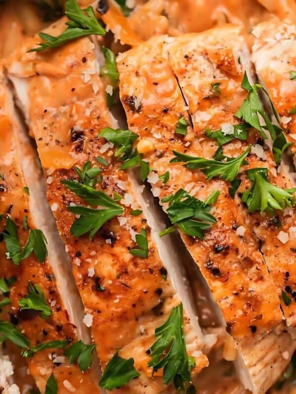 Close-up of sliced, seasoned chicken breasts garnished with fresh parsley and sprinkled with grated Parmesan cheese, served over creamy pasta with visible herbs.