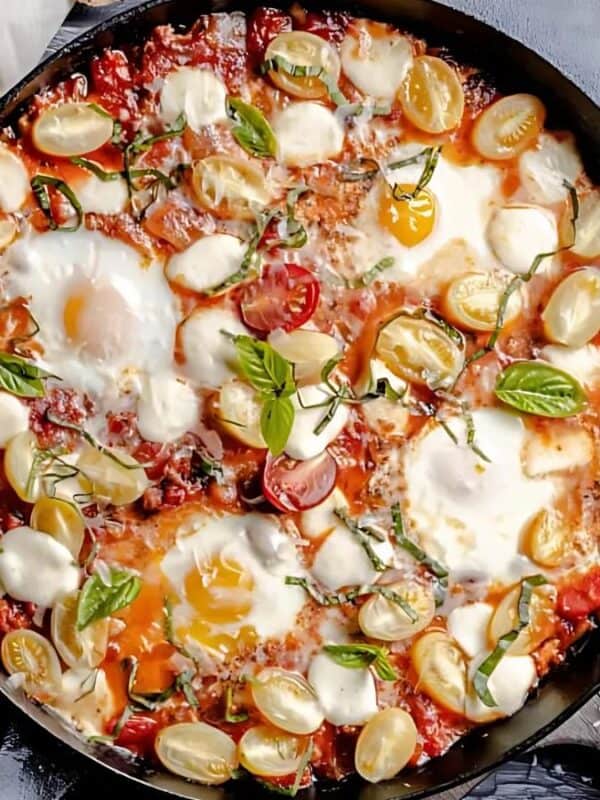 A skillet filled with shakshuka: poached eggs in a tomato sauce with cherry tomatoes, herbs, and melted mozzarella. Surrounding the skillet are a loaf of bread, a bowl of red pepper flakes, fresh basil, and cherry tomatoes.