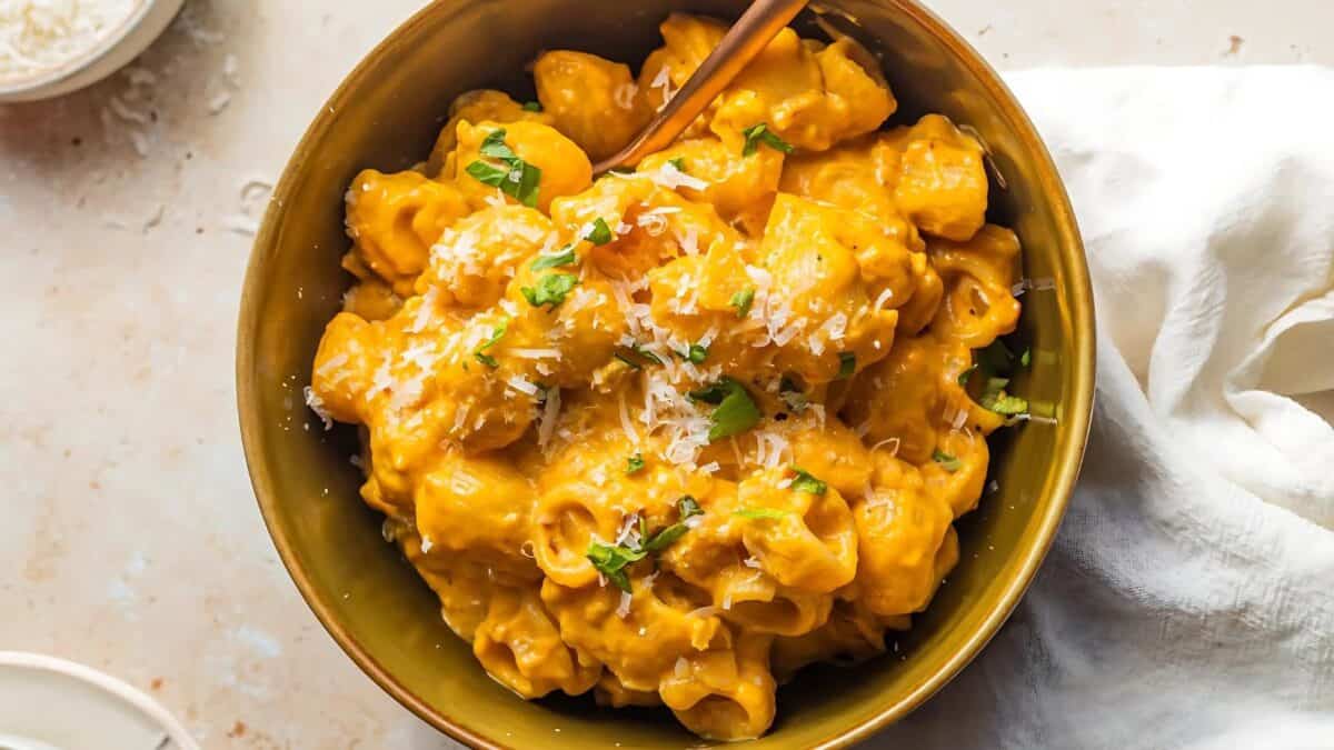 A bowl of creamy pumpkin pasta topped with grated cheese and garnished with fresh herbs. The pasta is served in a green bowl with a fork, placed on a light, textured surface with a white cloth napkin nearby.