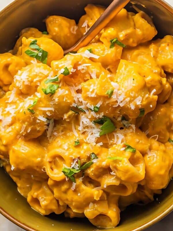 A bowl of creamy pumpkin pasta topped with grated cheese and garnished with fresh herbs. The pasta is served in a green bowl with a fork, placed on a light, textured surface with a white cloth napkin nearby.