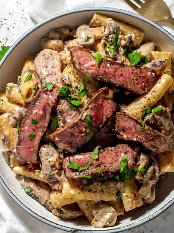 A bowl of creamy pasta topped with sliced steak, garnished with chopped parsley. The dish sits on a white cloth, next to gold utensils on a white speckled surface.