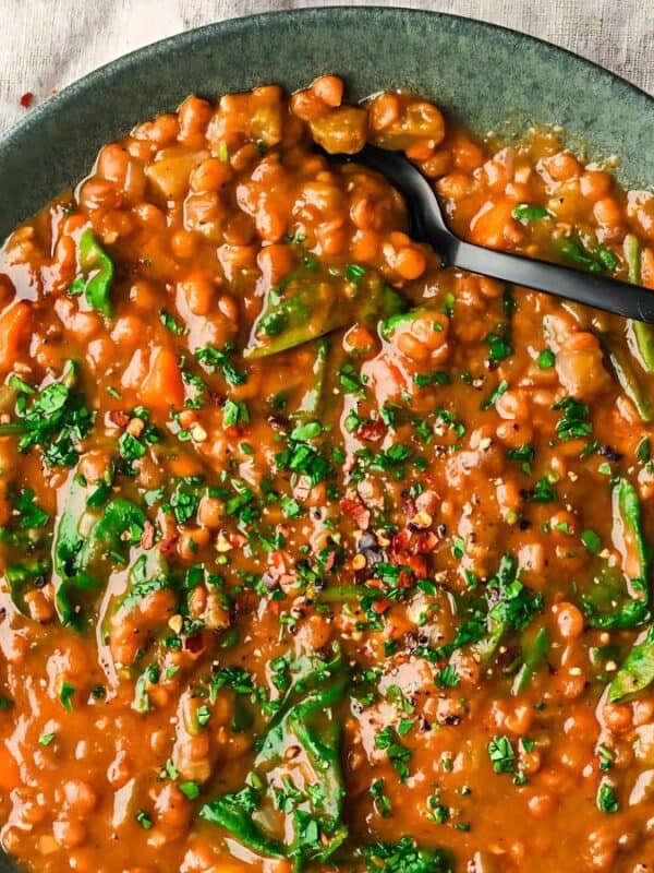 A green bowl filled with lentil stew topped with chopped parsley and red pepper flakes. The stew contains leafy greens and chickpeas. A black spoon rests on the bowl. The bowl is on a textured beige cloth. A glass is partially visible on the side.