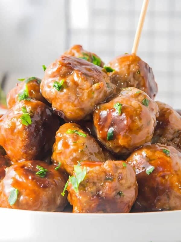 The image shows a white plate of meatballs in a brown sauce. The meatballs are arranged in a pyramid shape and are garnished with chopped parsley. Wooden skewers are stuck into several of the meatballs. A bowl of cherry tomatoes is in the background. A white and black striped napkin is in the lower left corner. The background is a white surface.