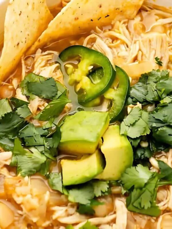 A white bowl filled with chicken and white bean soup, topped with sliced avocado, jalapeño, and cilantro. Tortilla chips garnish the side. A spoon rests in the bowl.