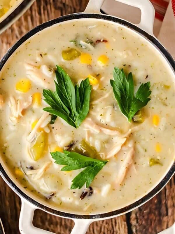 A bowl of creamy chicken and corn soup garnished with fresh parsley leaves. The soup is served in a white bowl on a wooden surface, accompanied by a red and white striped cloth. Other similar bowls are partially visible.
