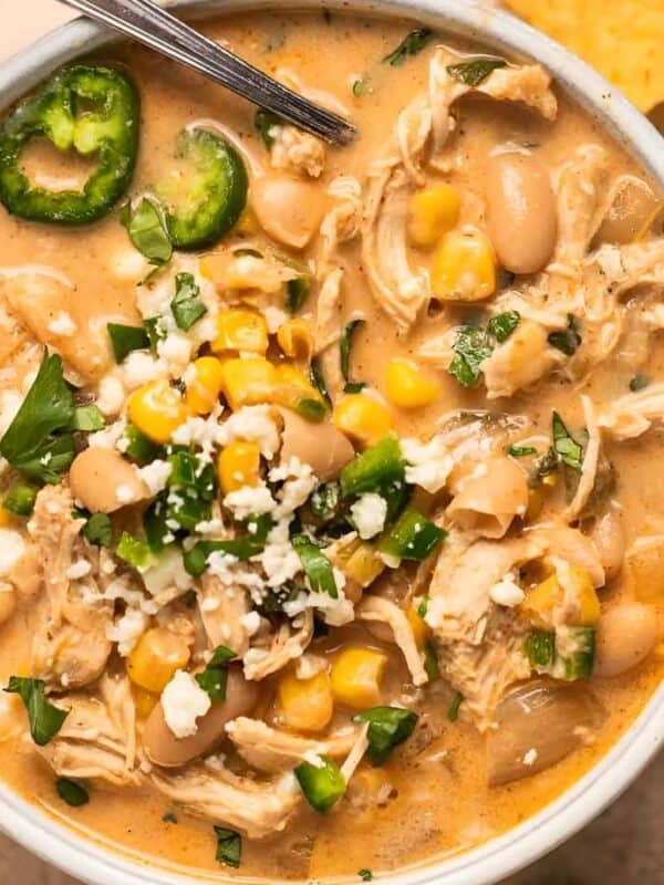 The image shows a bowl of creamy white chicken chili. The chili is topped with shredded cheese, chopped cilantro, sliced jalapeños, and a lime wedge. Tortilla chips are arranged around the bowl. The background is a light beige surface.