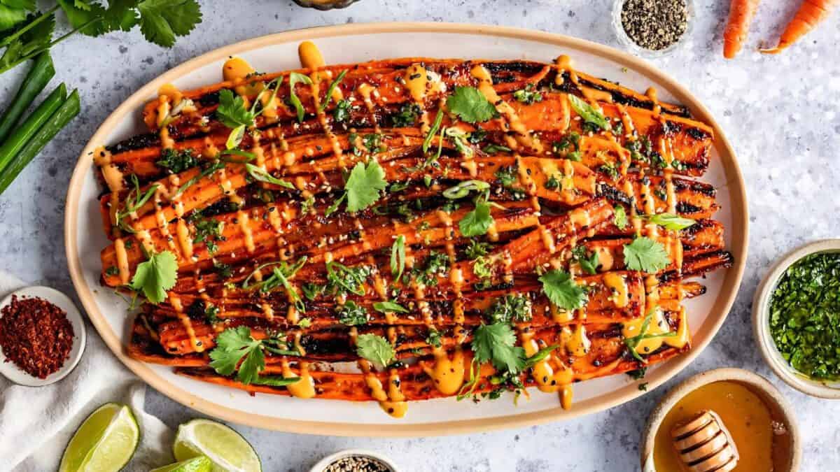 Grilled carrots drizzled with sauce, garnished with cilantro, on an oval platter. Surrounded by small bowls of spices, lime wedges, and a honey jar on a light surface. The presentation is vibrant and colorful.