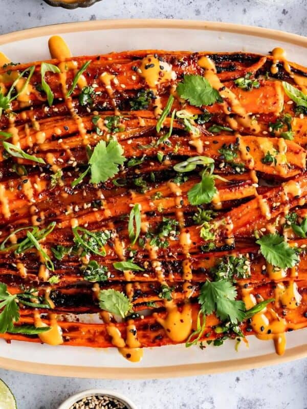 Grilled carrots drizzled with sauce, garnished with cilantro, on an oval platter. Surrounded by small bowls of spices, lime wedges, and a honey jar on a light surface. The presentation is vibrant and colorful.