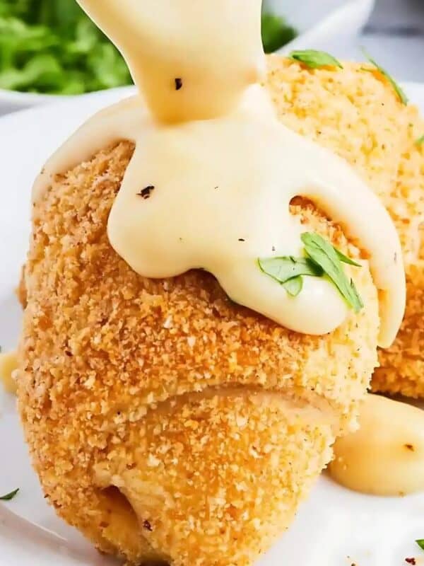 The image shows two breaded chicken rolls on a white plate. A creamy sauce is being poured over the rolls. Chopped parsley is sprinkled on the rolls and plate. The background is a blurry white surface with a bowl of parsley and some utensils.
