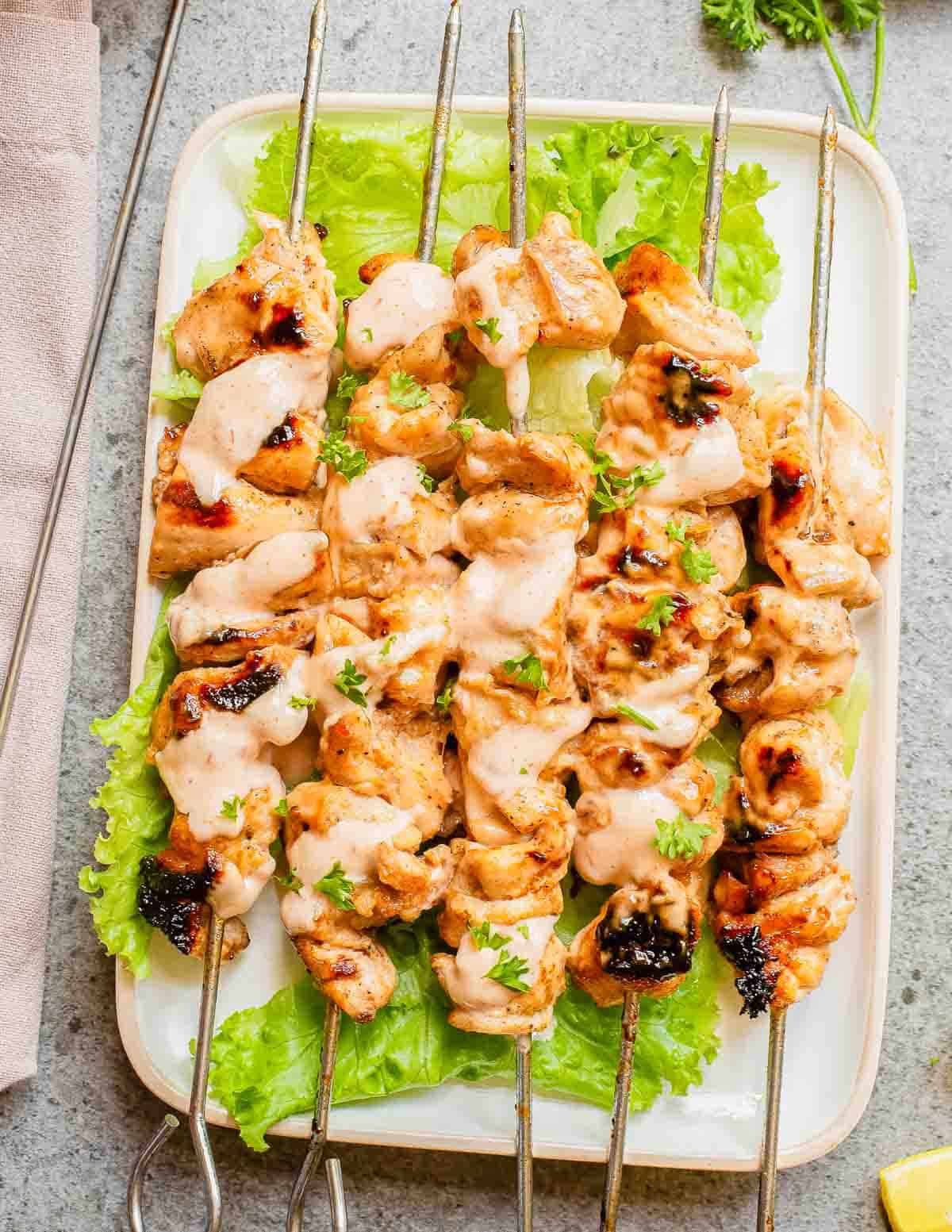 Bang bang chicken thigh skewers on a rectangular platter over lettuce with extra bang bang sauce drizzled on top.