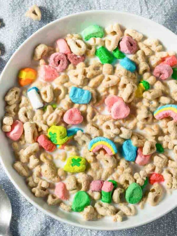 A bowl of colorful cereal with marshmallow shapes sits on a light blue fabric surface. The cereal features various marshmallow shapes, and scattered cereal pieces and two spoons are nearby.