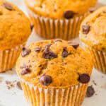 Four freshly baked muffins with chocolate chips on a white plate. The muffins have a golden-brown color and are topped with scattered chocolate chips. Some crumbs and chips are visible on the plate.
