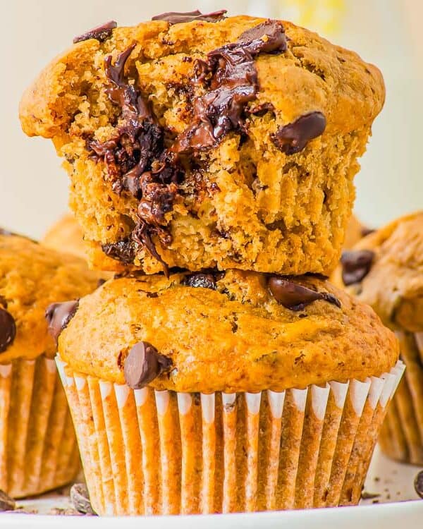Three peanut butter banana chocolate chip muffins are stacked on a plate. The top muffin has a bite taken out, revealing the moist interior with visible chocolate chips. Crumbs and scattered chocolate chips are visible on the plate.