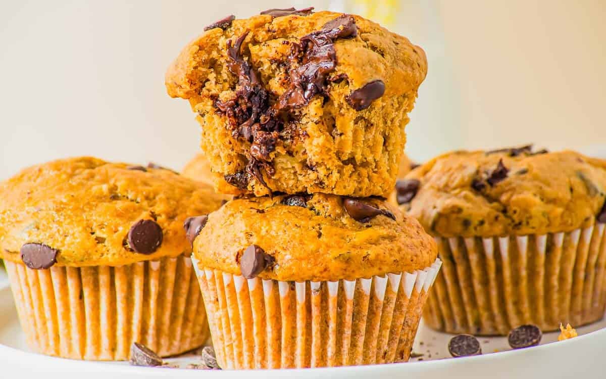 Three peanut butter banana chocolate chip muffins are stacked on a plate. The top muffin has a bite taken out, revealing the moist interior with visible chocolate chips. Crumbs and scattered chocolate chips are visible on the plate.