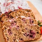 Sliced loaf of strawberry bread with a white glaze on top, resting on a wooden cutting board. The bread is visibly dense, with chunks of strawberries throughout. A few fresh strawberries are placed beside the loaf for garnish.