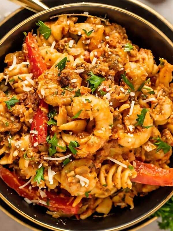 A bowl of pasta with shrimp, ground meat, red bell peppers, and herbs. The dish is garnished with grated cheese and fresh parsley. A small bowl of grated cheese is on the side.