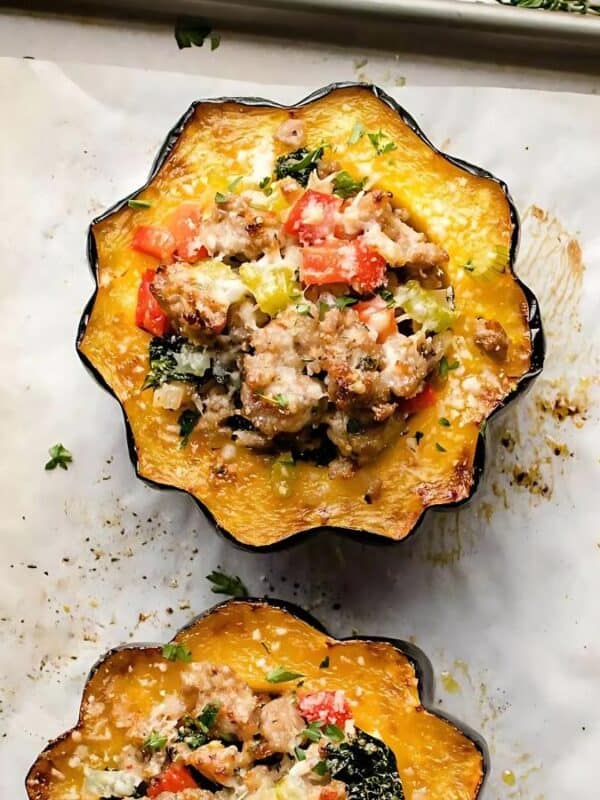 Stuffed acorn squash halves on a baking sheet. The squash is roasted with a filling of ground meat, diced tomatoes, leafy greens, and topped with melted cheese. Fresh herbs are sprinkled on top for garnish.