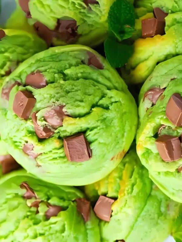 Several scoops of light green ice cream in a white bowl. The ice cream is studded with pieces of milk chocolate. Fresh mint leaves are visible around the bowl. The overall presentation is appetizing and suggests a refreshing and delicious dessert.