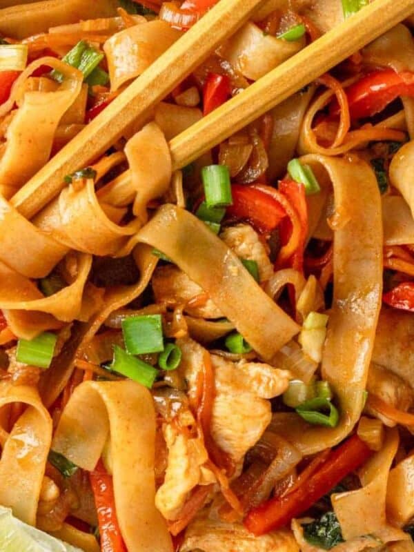 A bowl of wide rice noodles mixed with sliced chicken, red bell peppers, green onions, and basil. The dish is garnished with lime wedges and served with chopsticks. The noodles are coated in a brown, savory sauce.