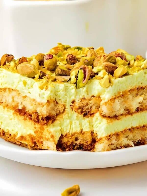 A slice of pistachio tiramisu on a white plate, topped with chopped pistachios. The dessert features visible layers of creamy filling and cake, set against a bright, clean background. Pistachios are scattered around the plate.