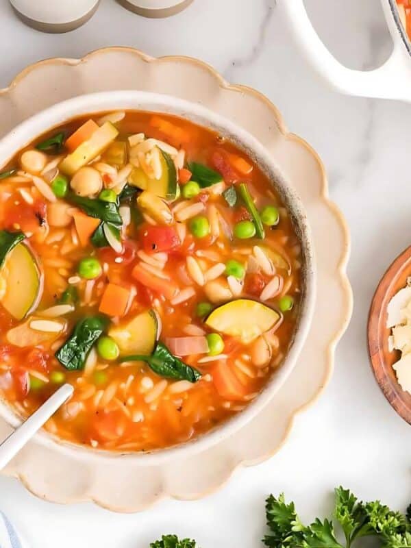 A bowl of vegetable soup with orzo, spinach, and zucchini sits on a white plate. Nearby are a pot of soup, a bowl of grated cheese, a striped napkin, and fresh parsley sprigs on a white table.