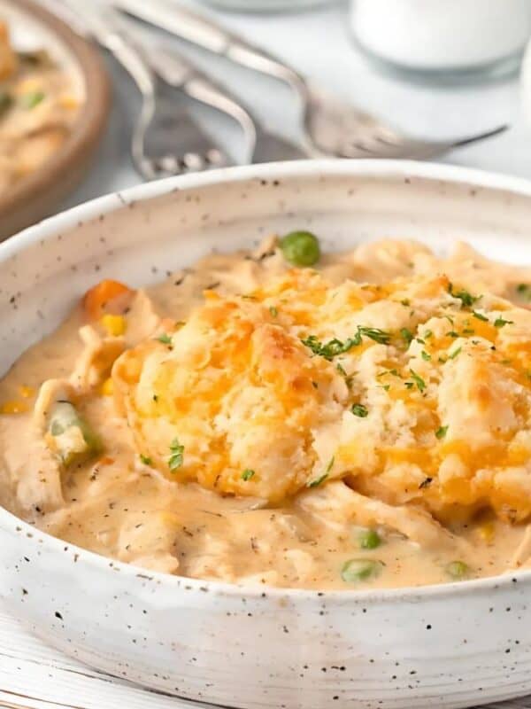 A rustic white bowl filled with creamy chicken pot pie stew, garnished with a golden biscuit on top. The stew includes visible peas, carrots, and chicken pieces. A gray cloth and utensils are in the background on a white wooden surface.