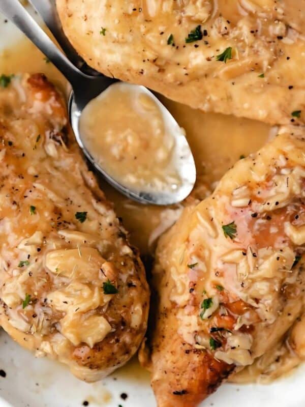 A white plate with three pieces of cooked chicken breasts covered in a creamy onion sauce. A silver spoon rests on top, and the dish is garnished with chopped herbs. A woven cloth is partially visible on the side.