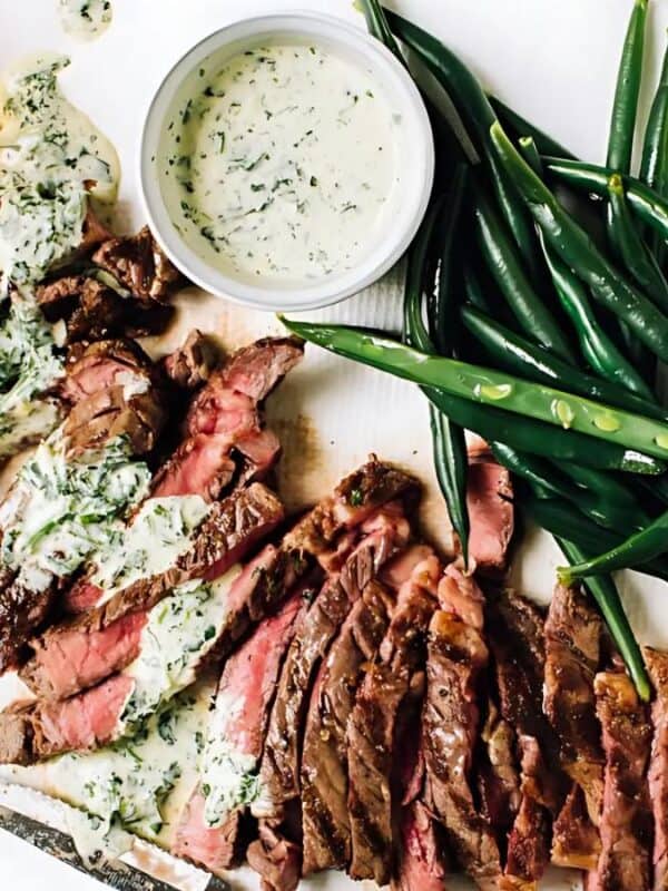 Sliced steak with herb sauce is arranged on a white plate alongside fresh green beans. A small bowl of additional sauce and a glass of beer are set beside the plate. The dish is presented on a white surface.