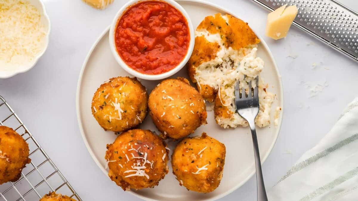 A plate of arancini, with crispy golden-brown rice balls, served alongside a bowl of marinara sauce. One arancini is cut open with a fork, revealing the cheesy interior. Grated cheese is sprinkled on top, with a grater and napkin nearby.