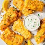 A plate of golden-brown beer battered fish pieces garnished with chopped herbs, accompanied by lemon wedges and a small bowl of tartar sauce. The dish is arranged on a white plate with a light-colored background.