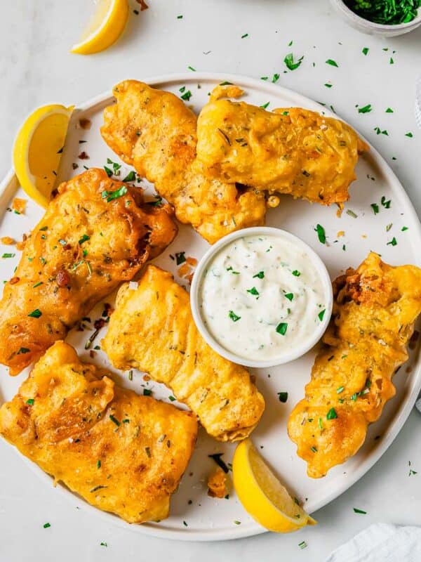 A plate of golden, crispy beer battered fried fish pieces garnished with chopped herbs and lemon wedges. A small bowl of creamy dipping sauce sits in the center. A glass of water and napkin are nearby on a light marble surface.