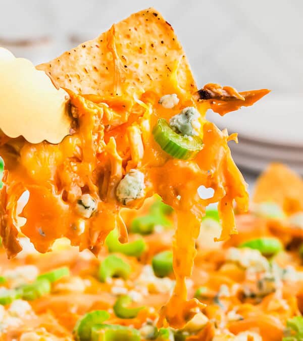 A close-up of a nacho being lifted with tongs. The nacho is covered in melted cheese, buffalo chicken, and topped with blue cheese crumbles and sliced celery. A blurred stack of plates can be seen in the background.