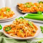 A plate of buffalo chicken nachos topped with melted cheese, sliced jalapeños, and chopped green onions. In the background, there's another plate of nachos and fresh celery sticks. The setting appears to be on a white surface.