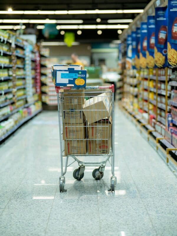 A shopping cart brimming with boxes sits in the center aisle of a grocery store, hinting at a savvy bulk buying strategy. Shelves packed with diverse products flank the shiny, clean floor, promising a successful hunt for all your food essentials.