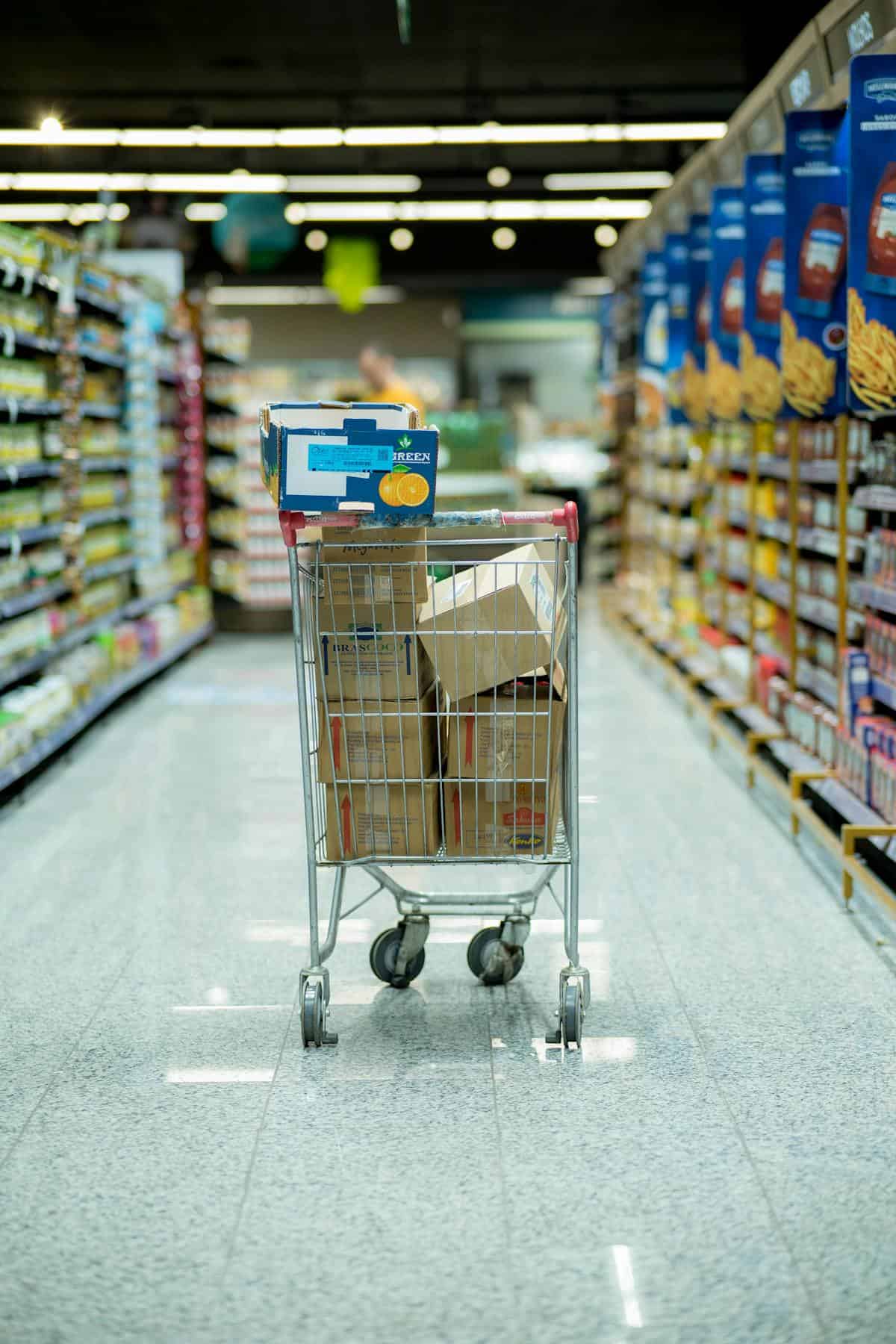 A shopping cart brimming with boxes sits in the center aisle of a grocery store, hinting at a savvy bulk buying strategy. Shelves packed with diverse products flank the shiny, clean floor, promising a successful hunt for all your food essentials.