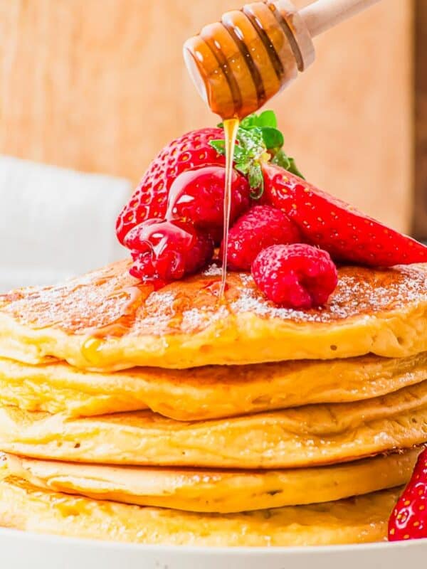 A stack of fluffy golden cottage cheese pancakes topped with fresh strawberries and raspberries on a white plate. Honey is being drizzled from a wooden dipper. A wooden board and a white cloth are in the blurred background.