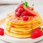 A stack of pancakes topped with strawberries, raspberries, and syrup on a white plate. A few strawberries and raspberries are scattered around the plate.
