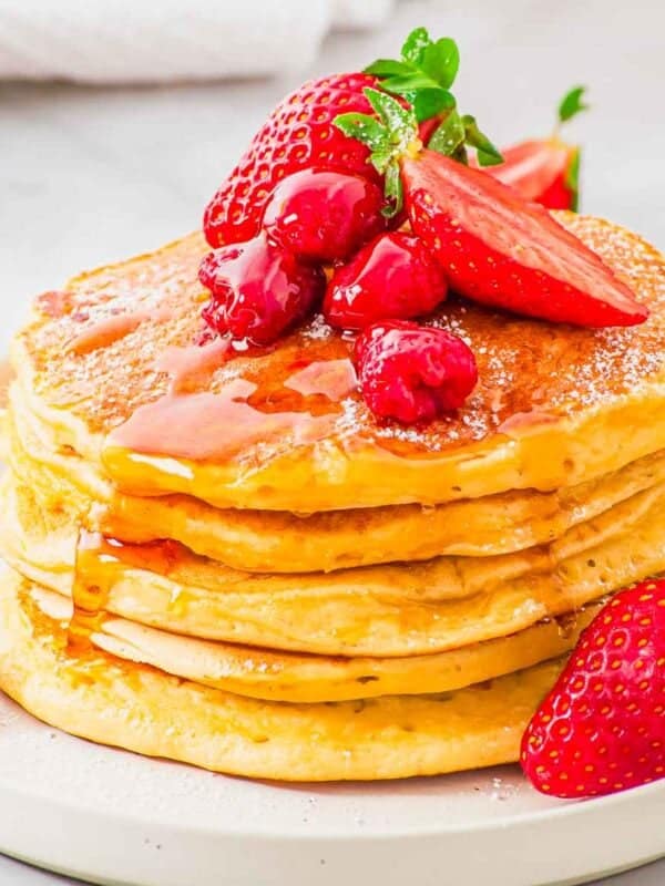 A stack of pancakes topped with strawberries, raspberries, and syrup on a white plate. A few strawberries and raspberries are scattered around the plate.