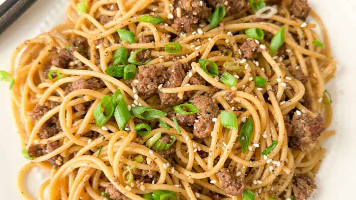A plate of spaghetti with ground beef, garnished with sliced green onions and sesame seeds. The dish is seasoned with visible black pepper.
