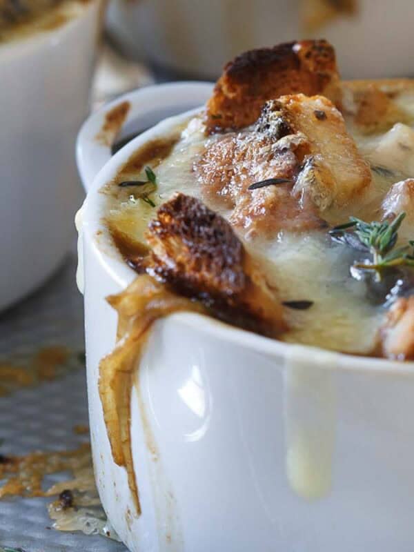 A close-up of a baked dish in a white ramekin, topped with melted cheese, toasted bread, and fresh thyme. The cheese has bubbled over the sides, and another similar dish is visible partially in the background. A spoon is adjacent to the dish.
