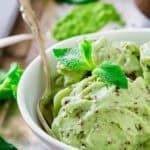 A bowl of matcha chocolate chip ice cream garnished with fresh mint leaves. The ice cream has visible chocolate flecks and is accompanied by a spoon. Surrounding the bowl are mint leaves and a small pile of matcha tea powder.