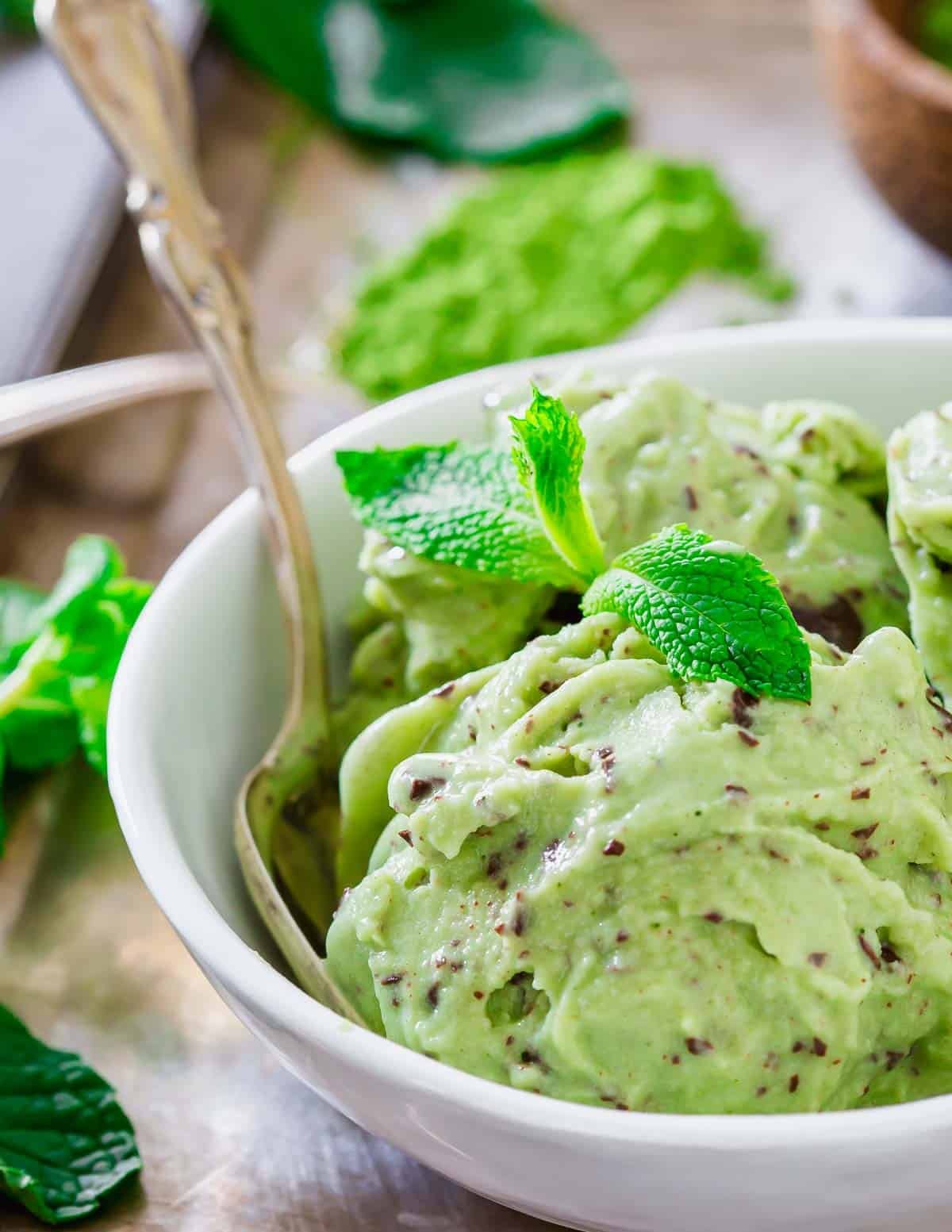 A bowl of matcha chocolate chip ice cream garnished with fresh mint leaves. The ice cream has visible chocolate flecks and is accompanied by a spoon. Surrounding the bowl are mint leaves and a small pile of matcha tea powder.